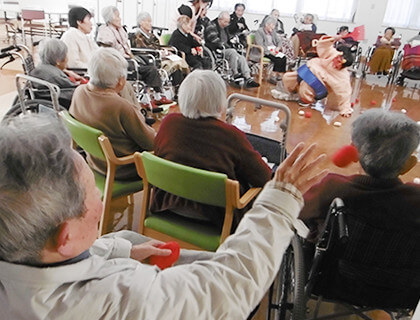 介護施設での集団レクリエーション。高齢者が輪になって楽器演奏や音楽活動に参加している様子。